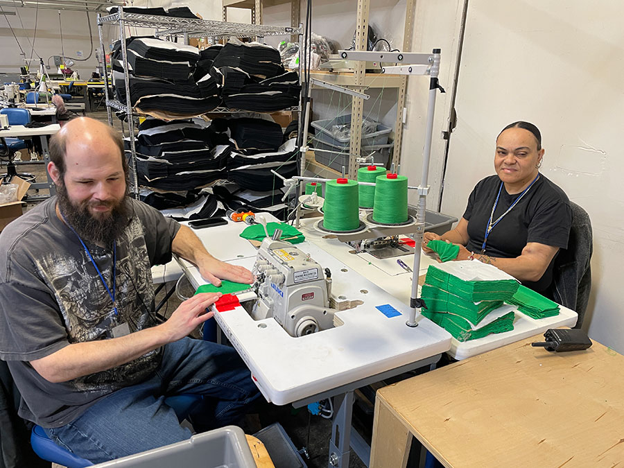 Two individuals working at Bestwork Industries using sewing machines in a workshop. Green thread spools and fabric pieces are visible on the tables. Shelves in the background contain stacked black fabric.