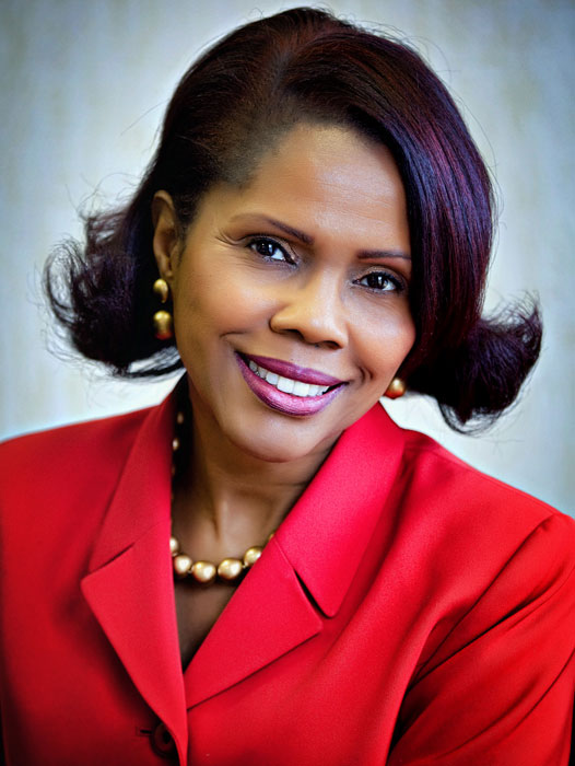 Woman with short, dark hair wearing a red blazer and pearl necklace, smiling against a neutral background