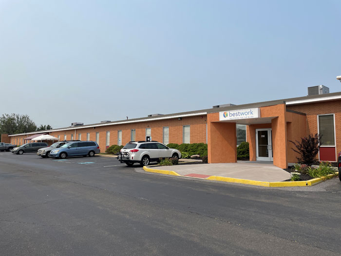 Bestwork Industries for the Blind building exterior, featuring a brick facade and a sign at the entrance. Several parked cars are visible in the parking lot.