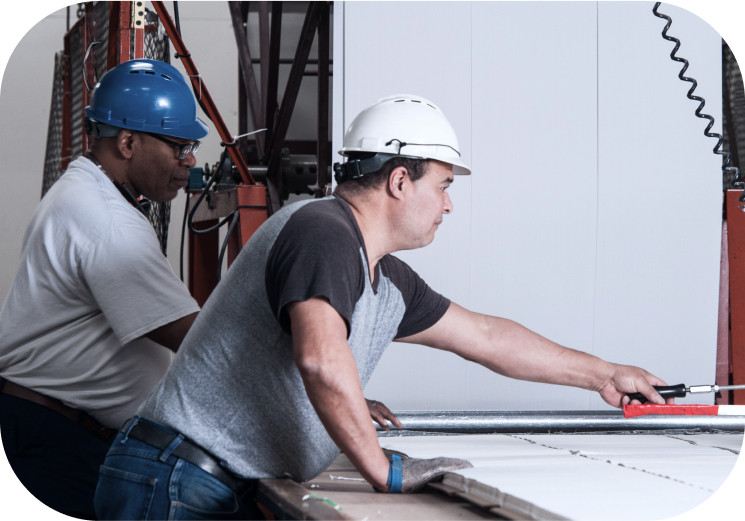 Two men working in a manufacturing setting, one wearing a blue hard hat and the other a white hard hat, operating machinery