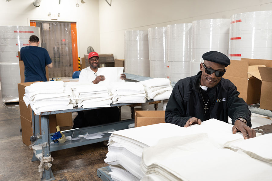 Warehouse setting with two men working. One man is folding white fabric while the other is standing behind a table. Stacks of fabric and cardboard boxes are visible in the background.