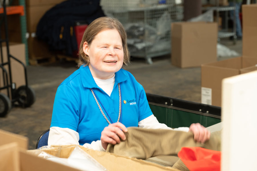 Woman in a blue shirt with bestwork industries, smiling while folding clothing in a warehouse setting with cardboard boxes.