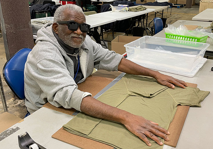 Man wearing sunglasses folding a green t-shirt at a table in a training facility for the blind