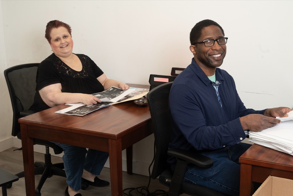 Office setting with one woman and one man working. One woman is seated at a desk with documents she's working to sort. The man is sorting papers. 