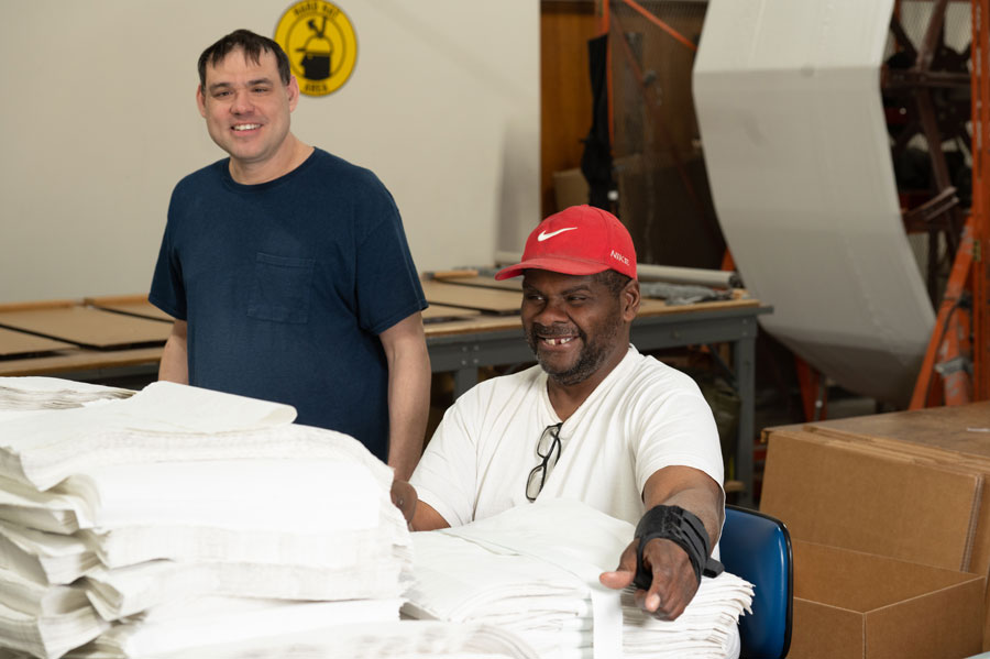 Bestwork Industries for the Blind training facility, two men smiling, one seated with stacks of white fabric