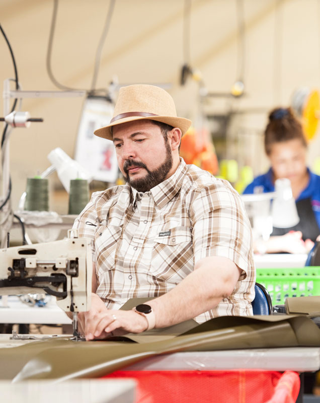 Man in a plaid shirt and hat sewing fabric at a workstation in a manufacturing facility. Another worker is visible