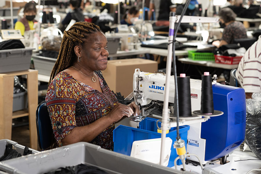 Sewing workshop with a woman operating a Juki sewing machine, surrounded by other workers and fabric bins