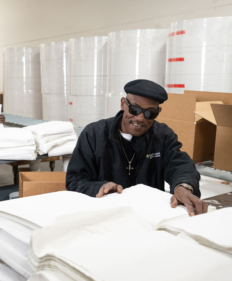 Warehouse setting with a man wearing sunglasses, sorting fabric sheets. Large rolls of material are stacked