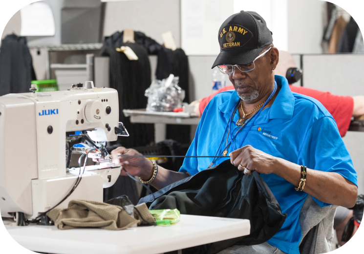 Man sewing at a JUKI sewing machine in a training facility, wearing a U.S. Army veteran cap and a blue shirt.