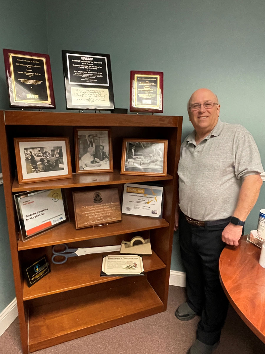 Wooden shelf displaying awards and photographs related to Bestwork Industries for the Blind, Inc. A man stands beside it