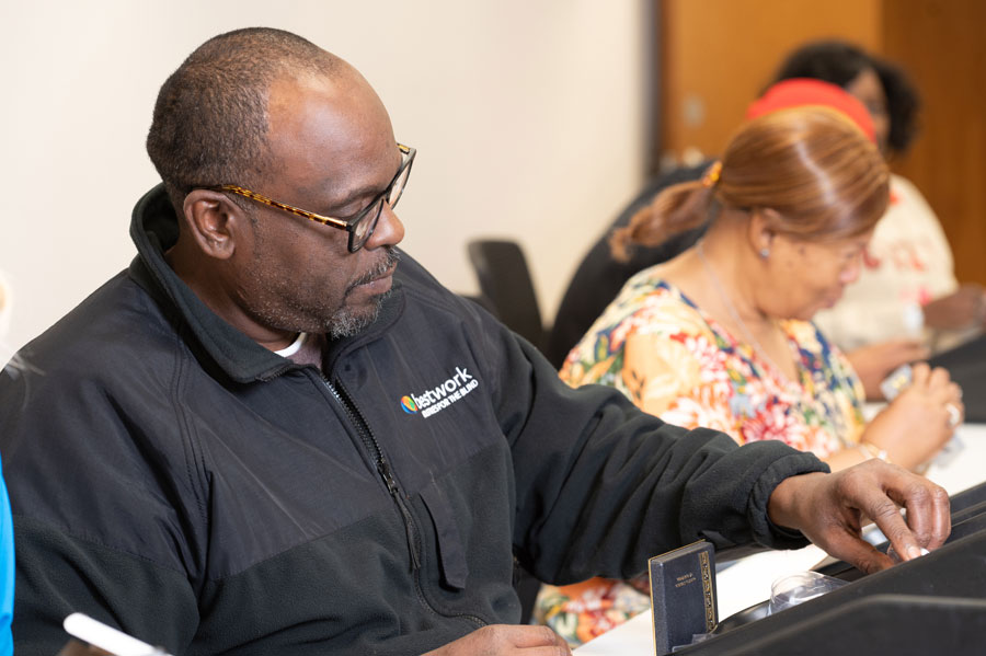 Training session at Bestwork Industries for the Blind; a man in a black jacket with the Bestwork logo is focused on a task, while a woman in a floral shirt is seated behind him.
