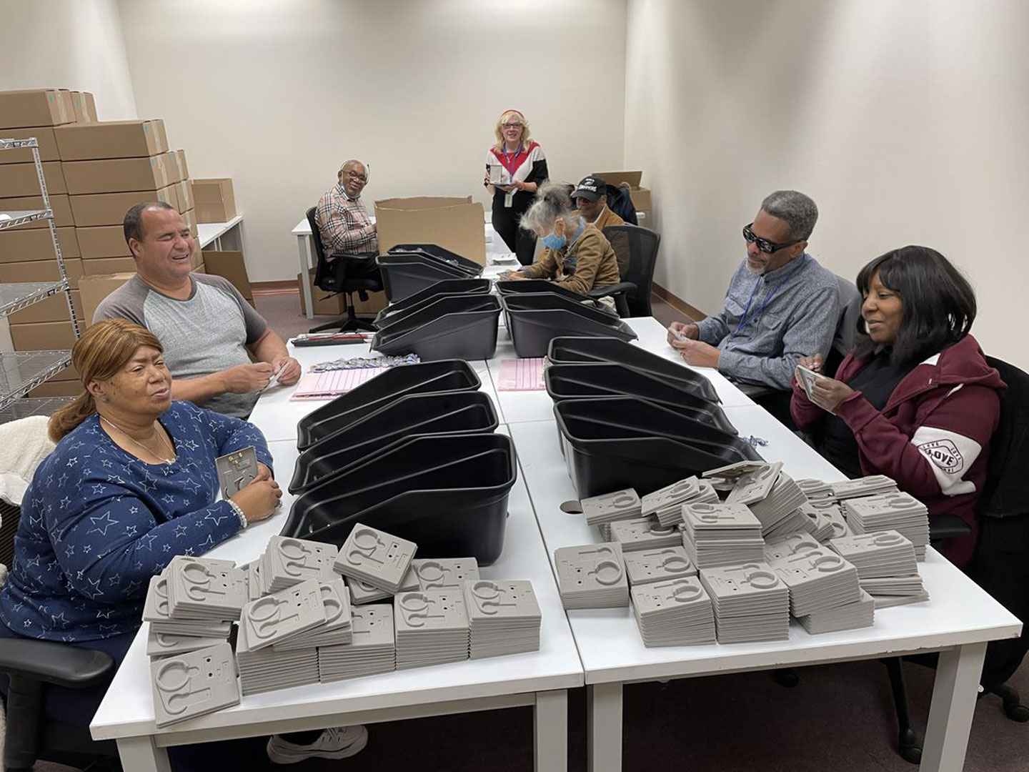 Training session at Bestwork Industries for the Blind, Inc. with eight participants seated at a table bins and stacks of gray products