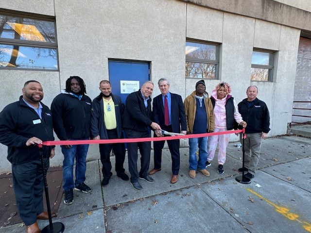 Ribbon-cutting ceremony outside Bestwork Industries for the Blind, Inc. building with eight individuals, including two holding scissors, in front of a door.