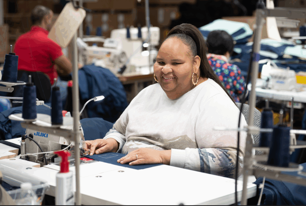 Woman smiling while working at a sewing machine in a workshop, surrounded by sewing equipment and fabric.