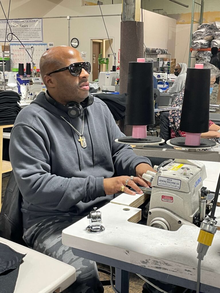 Man wearing sunglasses with headphones around his neck operates a sewing machine in a workshop setting with spools of black thread.