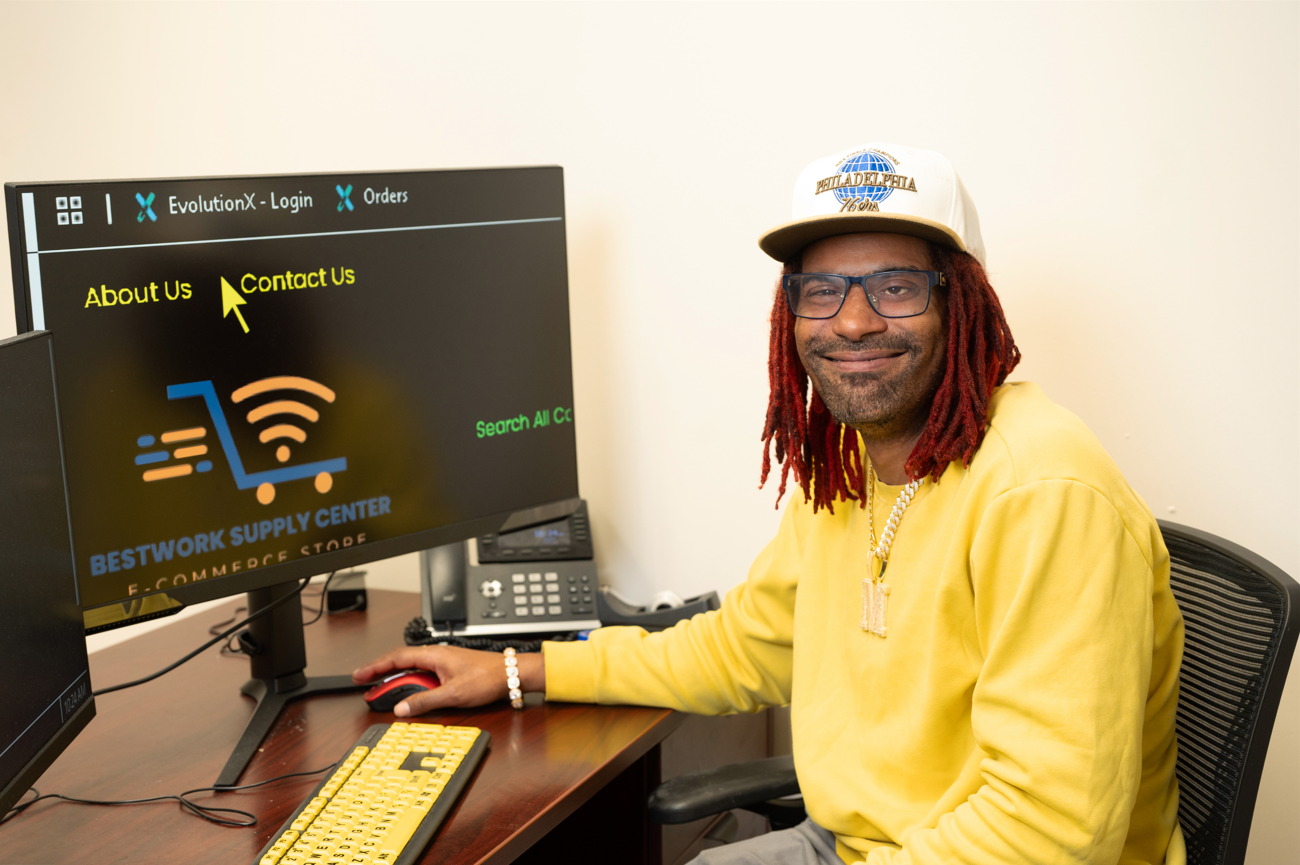 Man with red dreadlocks wearing a yellow sweatshirt and glasses sitting at a desk with a computer displaying the Bestwork Supply Center website.
