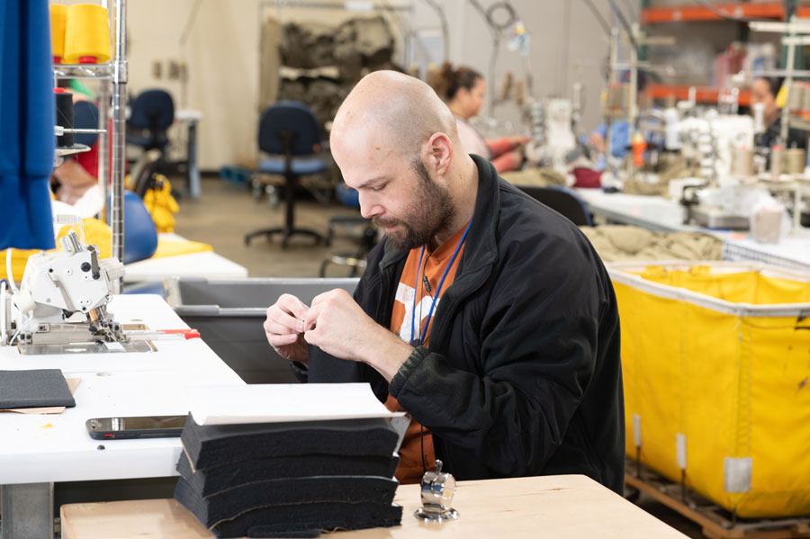 Workshop setting with a man working on a sewing project, surrounded by sewing machines and fabric stacks